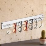 Transparante acryl brillenhouder aan een gestructureerde pleistermuur met zeven brillen, cactus op de plank eronder